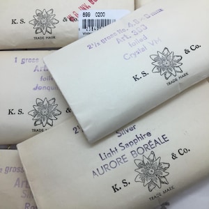May include: A collection of vintage paper packets, each with printed text and a floral logo. The packets are marked with details such as "Silver Starlight" and "Light Sapphire Aurore Boreale".