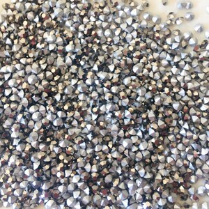 May include: A pile of small, faceted, silver-colored rhinestones. The rhinestones are scattered across the surface, catching the light and creating a sparkling effect. The gems have a multi-faceted cut, reflecting light from various angles.