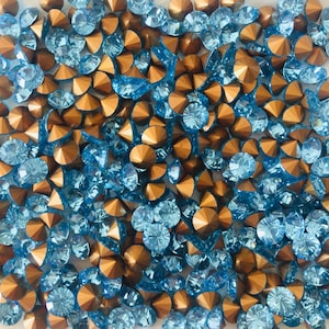 Könnte beinhalten: Eine Ansammlung von funkelnden, facettierten Strasssteinen in Hellblau und Kupferfarben. Die Edelsteine sind verstreut und erzeugen eine strukturierte, glitzernde Oberfläche. Ideal für Bastelarbeiten, Schmuckherstellung oder zum Verzieren.