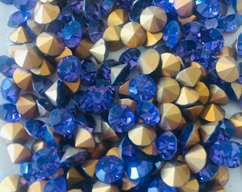 Può includere: Primo piano di strass dorati e blu scuro sparsi. Le pietre preziose sono sfaccettate, riflettendo la luce. La disposizione crea un effetto scintillante e strutturato, adatto per la creazione di gioielli o progetti artigianali. Le pietre sono misurate in millimetri.