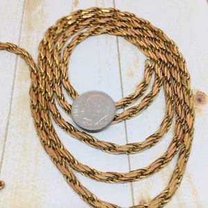 May include: A gold-toned chain necklace with a woven design, coiled on a light wood-grain surface. A U.S. dime is placed on the chain for size reference. The chain is made of small, interlocking links.