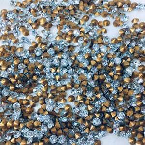 May include: A close-up shot displays a scattered collection of small, faceted rhinestones. The rhinestones are a mix of clear, silver-toned, and gold-colored, creating a sparkling effect. These embellishments are likely used for crafts, jewelry making, or garment decoration.