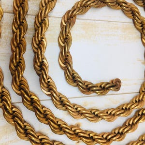 May include: A gold-colored rope chain necklace, coiled in a spiral pattern. The chain is made of tightly woven links, creating a textured surface. The necklace is displayed on a light-colored wooden surface.