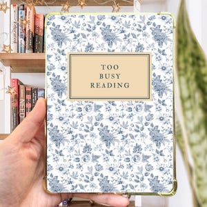 May include: A white and blue floral patterned iPad case with a gold trim. The case has a beige rectangle with the text "TOO BUSY READING" in black.