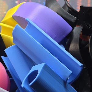 May include: A collection of colorful 3D-printed plastic parts in various shapes. The assortment includes blue, purple, yellow, black, pink, and green components. The parts appear to be for a construction or building set.