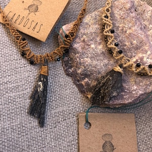 Może przedstawiać: Dwa brązowe naszyjniki z makramą z czarnymi koralikami i frędzlami z naturalnych włókien. Naszyjniki są prezentowane na brązowej, teksturowanej powierzchni z brązową papierową etykietą z napisem "CARDOSAS" i małą ilustracją kwiatu.