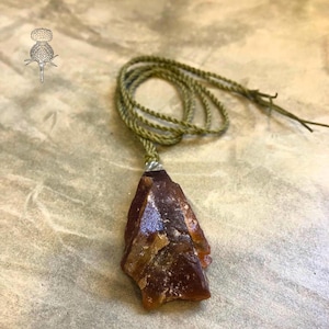 Op de afbeelding: Een bruine, onregelmatig gevormde hangerketting met een groen koord. De hanger is een ruwe, natuurlijke steen met een gepolijst oppervlak, dat licht reflecteert. Het koord is geknoopt en verstelbaar. De ketting is een uniek sieraad.