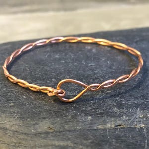 Può includere: Un braccialetto color rame con un design intrecciato. Il braccialetto è realizzato con fili sottili e intrecciati, che formano un anello continuo. La chiusura è una semplice chiusura a gancio e occhiello. Il braccialetto è esposto su una superficie grigio scuro.