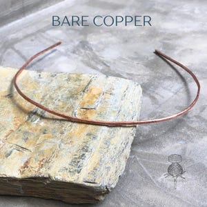 Op de afbeelding: Een blote koperen choker ketting met een gehamerde textuur, rustend op een ruwe, veelkleurige steen. De tekst "BARE COPPER" staat boven de ketting. De ketting is ongeveer 35.5 cm lang.