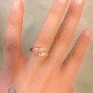 Peut inclure: Une bague en or rose enveloppante avec une surface texturée. La bague s'enroule autour du doigt avec un design ouvert. La bague est présentée sur une main.