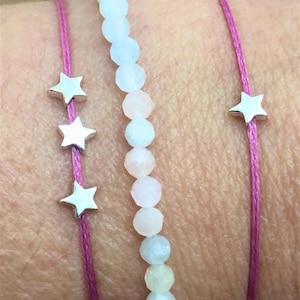 Pode incluir: Três pulseiras em um pulso. A pulseira superior é feita de contas azuis claras e rosa. A pulseira do meio é um cordão rosa com três pingentes de estrela prateados. A pulseira inferior é um cordão rosa com um pingente de estrela prateado.