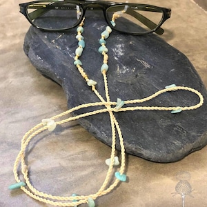 Può includere: Occhiali neri con una catena per occhiali con perline color crema con perline turchesi e color crema. Gli occhiali e la catena poggiano su una pietra blu scuro, un accessorio elegante.