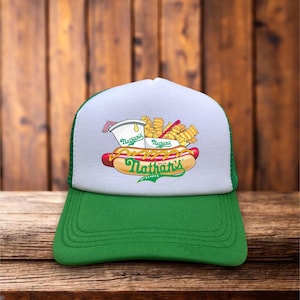 Pode incluir: Boné de caminhoneiro verde e branco com um gráfico de cachorro-quente, batatas fritas e uma bebida, com o texto "Nathan's" em verde e amarelo. O boné tem uma aba verde e uma parte traseira de malha.