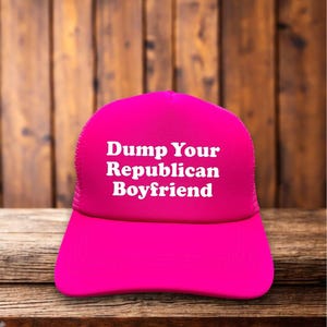 May include: A bright pink trucker hat with the text "Dump Your Republican Boyfriend" in white block letters. The hat has a mesh back and a curved brim, set against a wooden background.