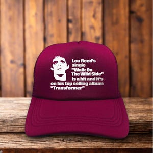 Può includere: Cappellino da camionista bordeaux con grafica bianca di Lou Reed e testo sulla sua canzone "Walk on the Wild Side" dall'album "Transformer". Il cappellino ha un retro in rete.