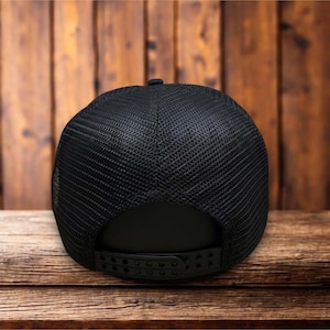 Puede incluir: Gorra de camionero negra con parte trasera de malla y visera curva. La gorra tiene un cierre a presi&oacute;n para ajustar la talla. La gorra se muestra sobre una superficie de madera.