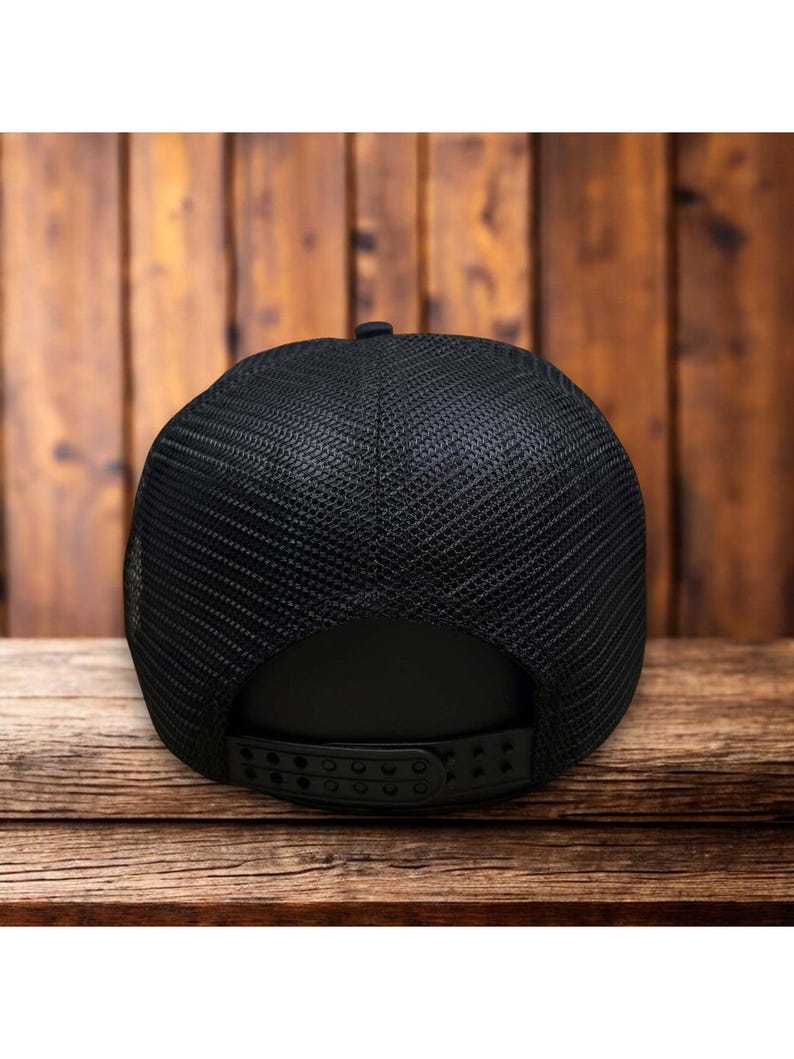 Puede incluir: Gorra de camionero negra con parte trasera de malla y visera curva. La gorra tiene un cierre a presi&oacute;n para ajustar la talla. La gorra se muestra sobre una superficie de madera.