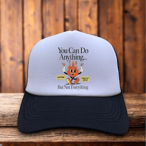 May include: A black and white trucker hat with a light grey front panel. The panel features the text "You Can Do Anything... But Not Everything" and a cartoon flame character. The hat has a curved peak.