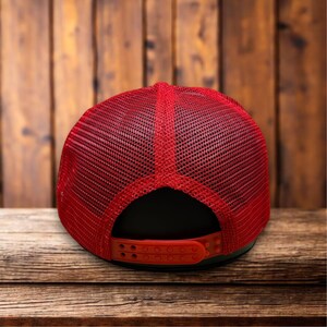 Puede incluir: Gorra de camionero roja con la parte trasera de malla y visera curva. La gorra tiene un panel frontal negro y un cierre a presi&oacute;n rojo ajustable. La gorra est&aacute; sobre una superficie de madera.