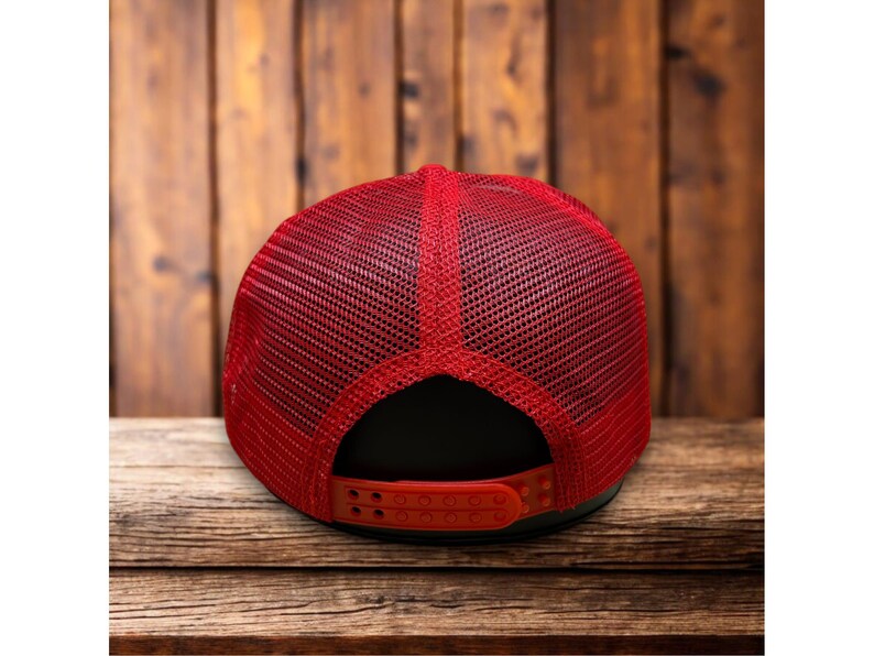 Puede incluir: Gorra de camionero roja con la parte trasera de malla y visera curva. La gorra tiene un panel frontal negro y un cierre a presi&oacute;n rojo ajustable. La gorra est&aacute; sobre una superficie de madera.