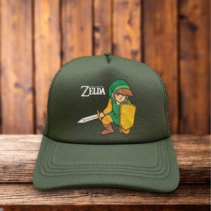 May include: Olive green trucker hat featuring a cartoon illustration of a character from The Legend of Zelda video game. The character is depicted with a sword and shield. The text "ZELDA" is above the character.
