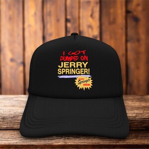 Puede incluir: Gorra de camionero negra con el texto "I GOT DUMPED ON JERRY SPRINGER!" en rojo y amarillo. La gorra también presenta las palabras "Jerry Springer show" en amarillo sobre una estrella.