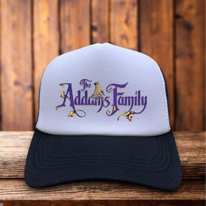 May include: A black and white trucker hat with a white front panel. The front features the text "The Addams Family" in purple script with a hand graphic. The hat is on a wooden surface.