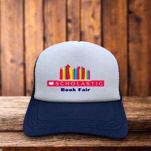 Peut inclure: Casquette de camionneur bleu marine et blanche avec l'inscription "Scholastic Book Fair" et une illustration de livres colorés. Le chapeau est posé sur une surface en bois.