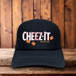 May include: Black trucker hat with the text "CHEEZ-IT" in white letters with a pink checkered background. The hat also features the words "Baked Snack Crackers" and images of the crackers.