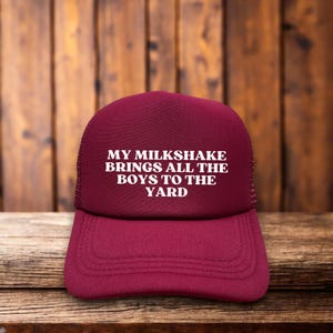 Könnte beinhalten: Eine burgunderfarbene Trucker-Kappe mit weißem Netzrücken. Die Vorderseite der Kappe trägt den Text "MY MILKSHAKE BRINGS ALL THE BOYS TO THE YARD" in Weiß.
