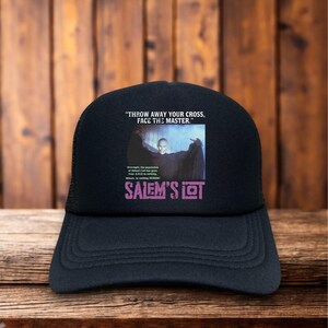May include: Black trucker hat with a mesh back and a curved brim. The front features a graphic from "Salem's Lot" with the text "THROW AWAY YOUR CROSS, FACE THE MASTER." and the title "SALEM'S LOT" in purple.