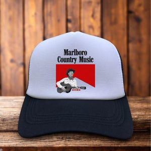 Może przedstawiać: Czarno-biała czapka typu trucker z białym panelem przednim. Na czapce widnieje napis "Marlboro Country Music" nad grafiką kowboja grającego na gitarze na czerwonym tle.