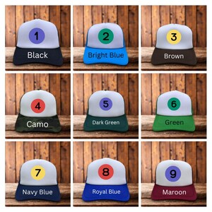 Puede incluir: Una colecci&oacute;n de nueve gorras de camionero en varios colores, cada una con un panel frontal blanco y una visera de color. Las gorras est&aacute;n etiquetadas con n&uacute;meros y nombres de colores: Negro, Azul brillante, Marr&oacute;n, Camuflaje, Verde oscuro, Verde, Azul marino, Azul real y Granate.