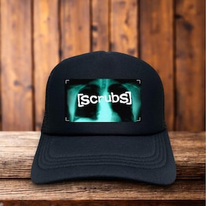 May include: Black trucker hat with a curved brim and mesh back. The front panel features a rectangular graphic of a chest X-ray with the word "scrubs" in white text. The hat is set against a wooden background.