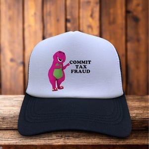 May include: A black and white trucker hat with a white front panel featuring a cartoon purple dinosaur with a green belly. The text "COMMIT TAX FRAUD" is printed in black.