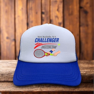 May include: A blue and white Challenger tennis cap with a mesh back. The front features the text "New Rochelle, N.Y. CHALLENGER" with a tennis racket and ball graphic. The cap is on a wooden surface.