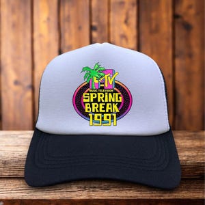 May include: A black and white trucker hat with a white front panel featuring the MTV Spring Break 1991 logo. The logo includes a palm tree, the MTV logo, and the words "Spring Break 1991" in a colorful, retro design.