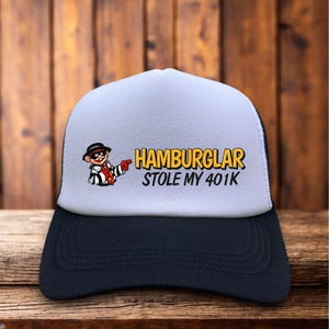 May include: A trucker hat with a white front panel and a black brim. The hat features a cartoon image of a Hamburglar character and the text "HAMBURGLAR STOLE MY 401K" in yellow and black.