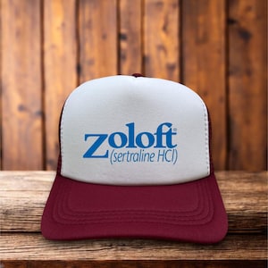 May include: A burgundy and white trucker hat with the word "Zoloft" in blue script across the front. Below the word "Zoloft" is the text "(sertraline HCI)". The hat has a burgundy brim and mesh backing.