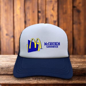 May include: A navy blue and white trucker hat with the text "McChicken Sandwich" and the McDonald's logo. The hat has a mesh back and a curved brim. The logo is yellow and blue.