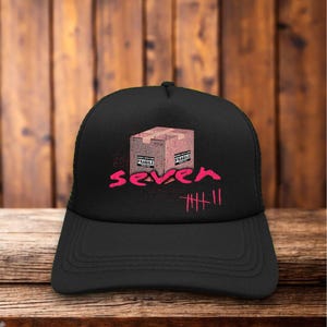 Könnte beinhalten: Schwarze Trucker-Cap mit einem rosa Aufdruck einer Pappschachtel mit der Aufschrift "FRAGILE" und dem Wort "seven" in Rosa. Unter "seven" befinden sich fünf vertikale Striche. Die Kappe hat einen Mesh-Rücken und einen gebogenen Schirm.