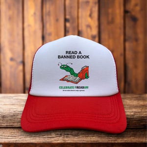 May include: A red and white trucker hat with the text "READ A BANNED BOOK" and a cartoon worm reading a book. The hat also features the text "CELEBRATE FREEDOM" in green.