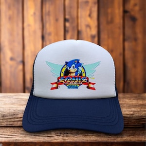 May include: A navy blue and white trucker hat featuring the Sonic the Hedgehog logo. The logo includes a blue hedgehog character with wings, the word "SONIC", and the SEGA logo. The hat has a mesh back.