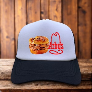 Pode incluir: Boné de caminhoneiro com painel frontal branco e parte traseira de malha preta. A frente apresenta um gráfico de um sanduíche Arby's e o logotipo da Arby's em vermelho. O boné está em uma superfície de madeira.