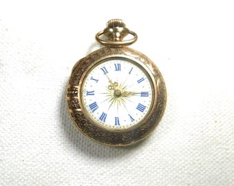 May include: A gold-toned pocket watch with a white face and blue Roman numerals. The watch has an ornate, engraved bezel and a small loop at the top. The hands are gold, and the watch face has a starburst design.