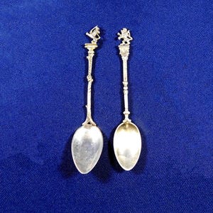 May include: Two ornate silver spoons with detailed handles and oval bowls. The handles feature decorative elements, including what appears to be a lion or griffin at the top. The spoons are set against a deep blue background.