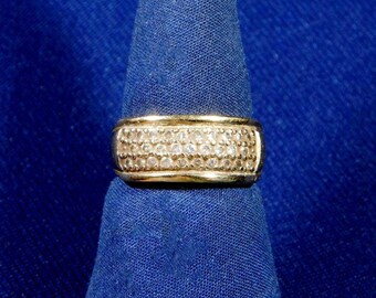 May include: A gold-toned ring with a wide band, densely set with small, clear gemstones. The ring is displayed against a deep blue fabric background, highlighting its sparkle and design.