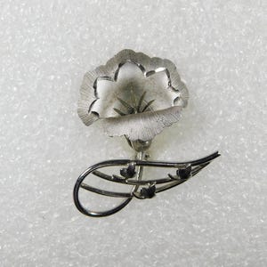 May include: Silver tone flower brooch with a stylized flower design and leaves. The flower has a textured surface and the leaves are delicate and intricate.