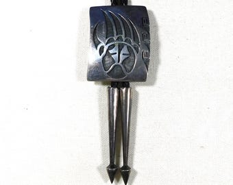 May include: Silver bolo tie with a rectangular pendant featuring a bear claw design. The pendant is connected to a black braided cord and has two silver-toned pointed tips. The bolo tie is set against a white background.