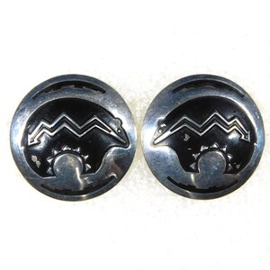 May include: Pair of silver-toned round stud earrings. Each earring features a black bear design with a zigzag pattern and a sun-like element. The earrings have a detailed, vintage aesthetic.